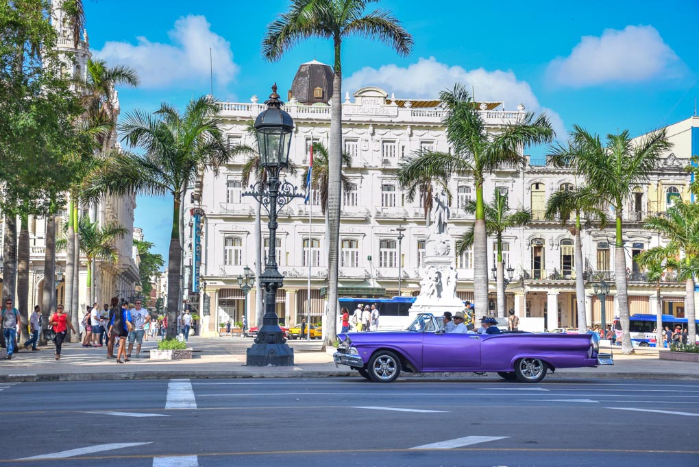 An old car sits at the lights in Cuba with beautiful buildings in the background