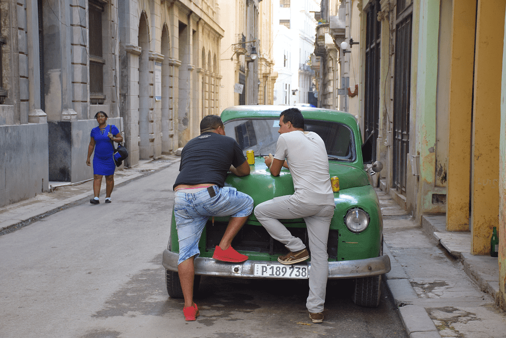 Two Cubans share a beer in the street on a vintage car