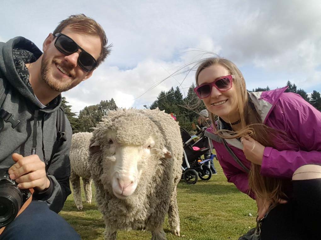 Bailey and Daniel from Destinationless Travel pose for a photo with a sheep in New Zealand
