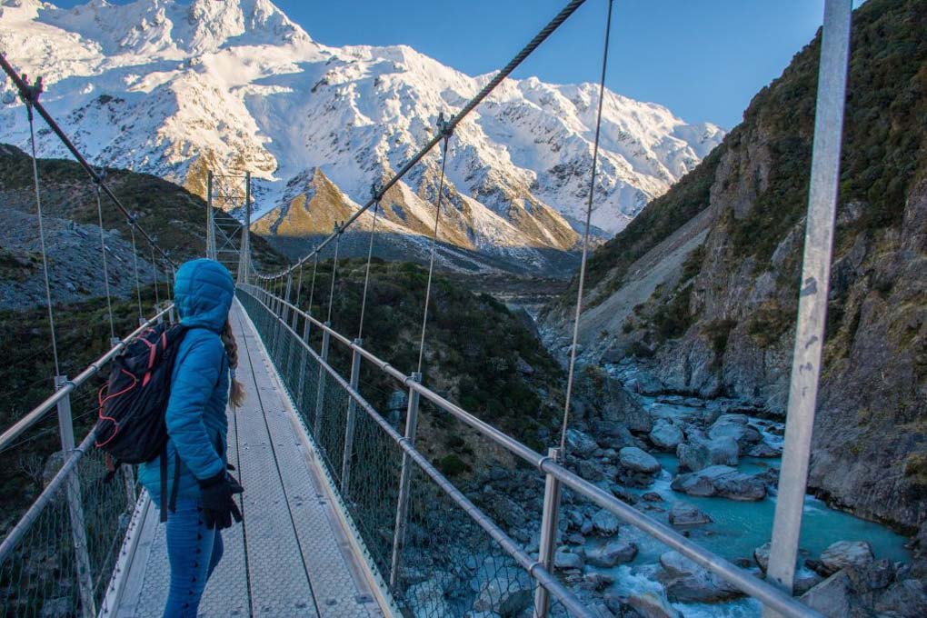 A woman stands on the Hooker Valley track suspension bridge looking towards Mt Cook