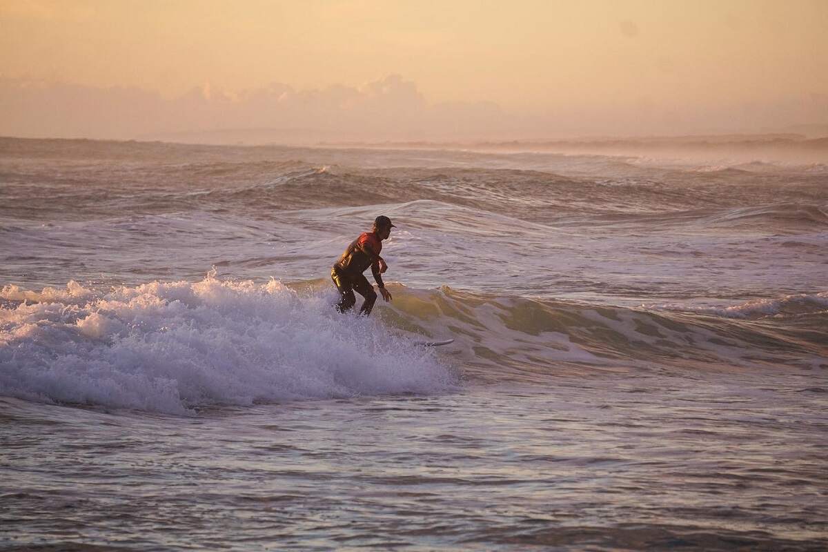 a man surf at sunset at Muriwai Beach, a pink sky behind him.