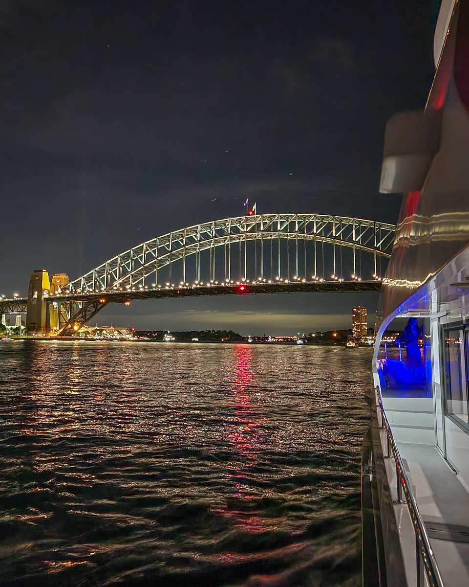 A boat cruises under towards the Syndey Harbour bridge at night during a dinner cruise