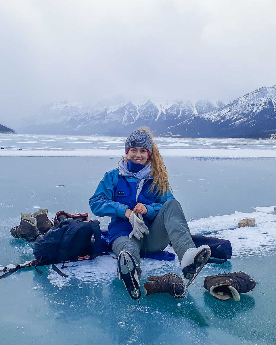 Bailey sits on the ice on Abraham Lake, lacing up her skates with mountains behind her