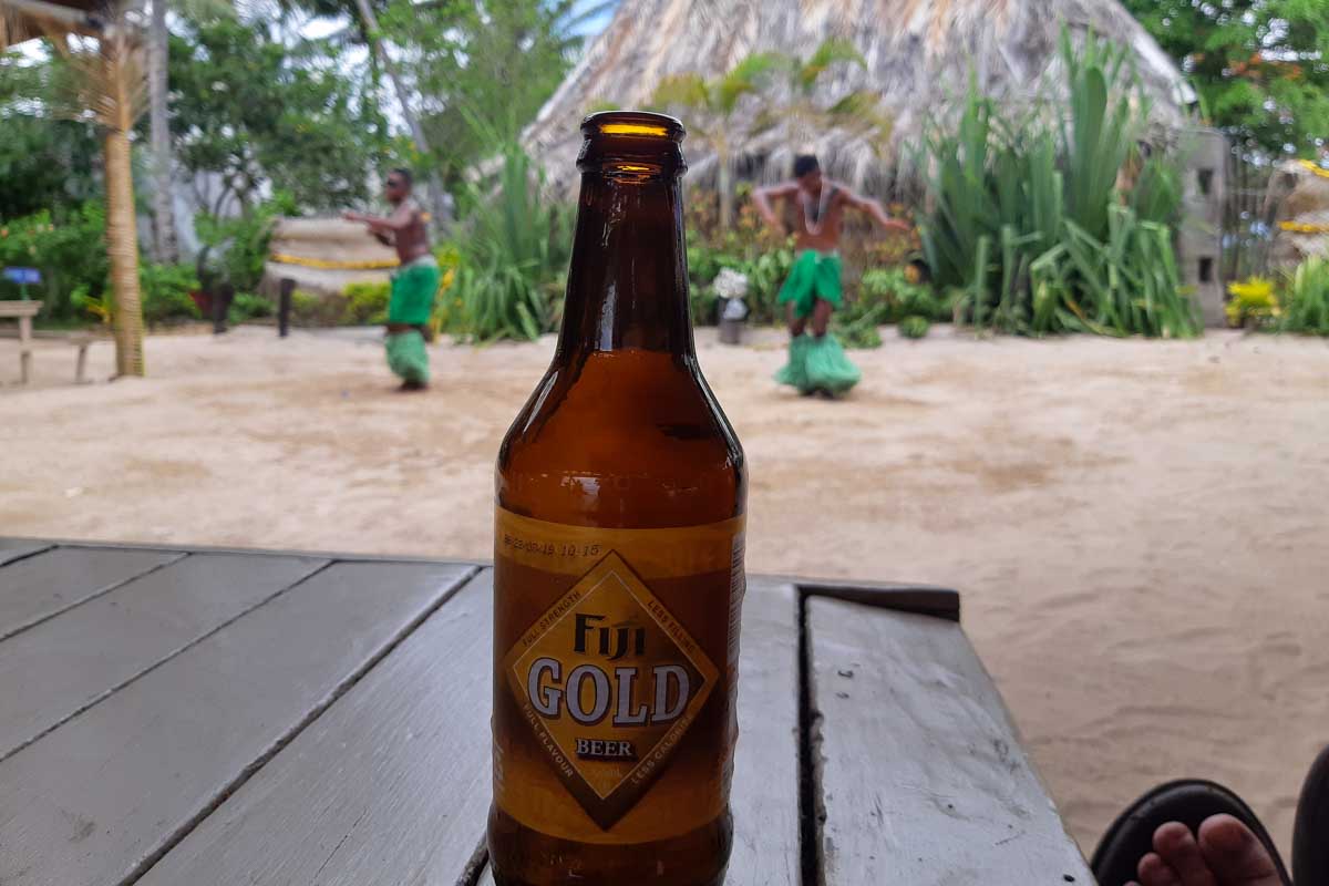 The local beer fiji gold on a table as fijians perform a dance in the background robinson crusoe island