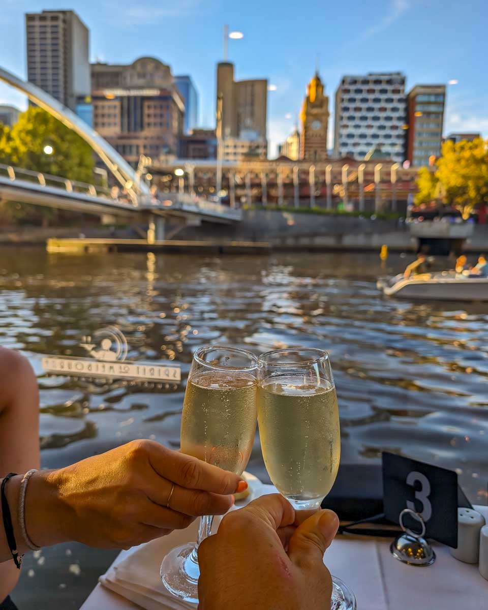 Bailey and Daniel cheers their wine on the spirit of melbourne dinner cruise with tthe city in the background